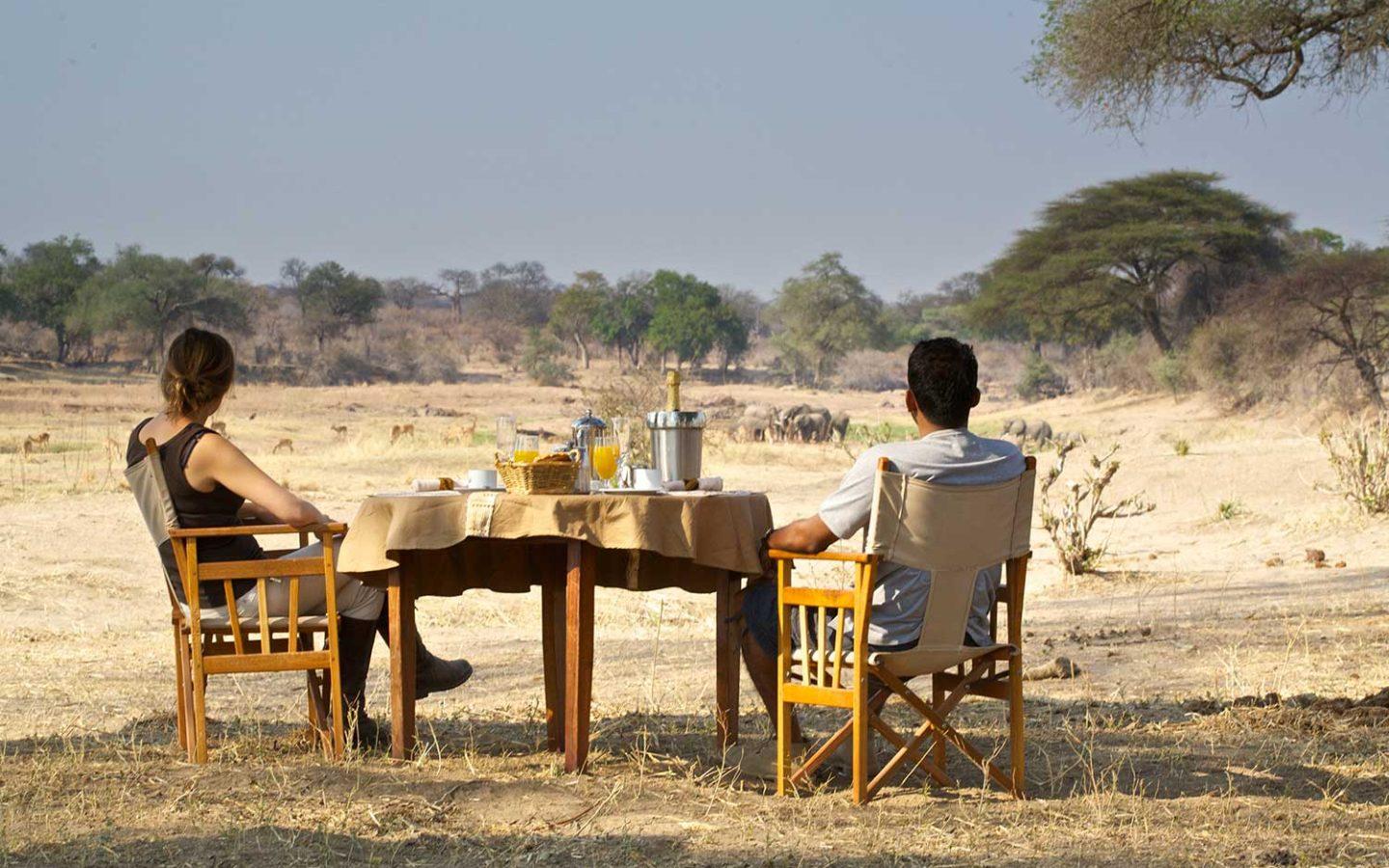 a man and woman sit on wooden chairs with lunch table while in the middle of